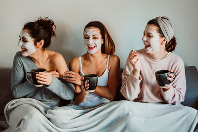 Cheerful female friends with facial masks holding mugs while sitting against wall at home