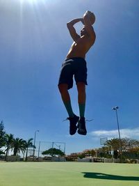 Low angle view of shirtless man playing basketball on field against clear sky during sunny day
