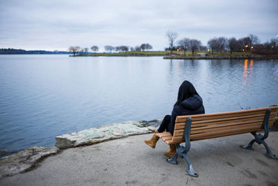 Rear view of woman sitting on seat by lake against sky