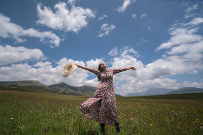 Woman alone with flowered dress and hat raising her arms and head in field during a sunny morning