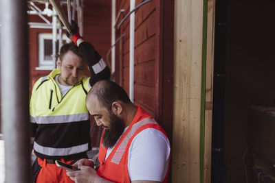 Male construction worker looking at bearded colleague using smart phone at site