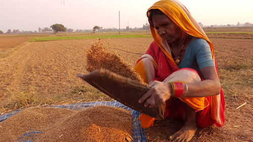 Mature woman winnowing crops in basket while crouching on field