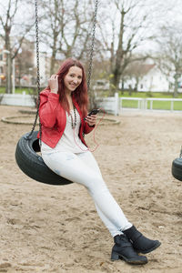 Full length of happy teenage girl enjoying music on tire swing in playground