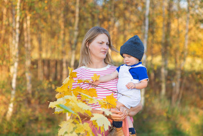Woman holding baby boy against trees during autumn