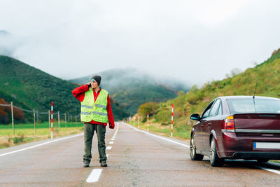 Female driver in reflective vest standing on road near broken car in mountainous area and talking on smartphone while calling for help