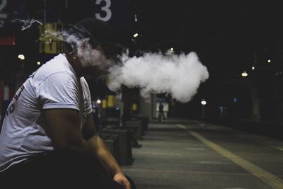 Side view of man exhaling smoke while sitting at railroad station platform