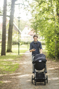 Man using smart phone while pushing baby carriage on footpath at public park