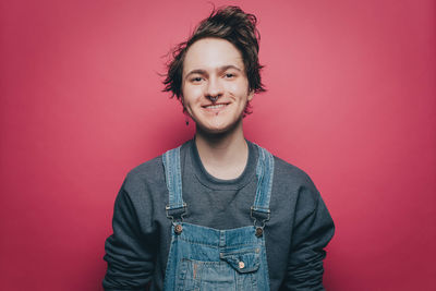 Portrait of smiling young man wearing denim overalls over pink background