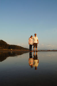 Couple standing at lakeshore against sky during sunset