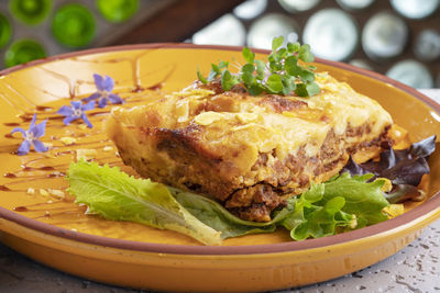 Lasagna on an orange plate accompanied by lettuce leaves, watercress and purple flowers.