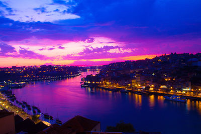 Illuminated city by river against sky at sunset