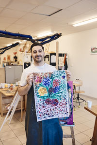 Portrait of smiling male painter showing multi colored canvas painting in art class