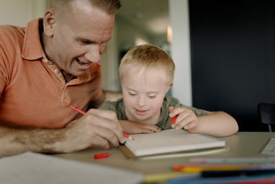 Father assisting son with down syndrome in writing on book at dining table