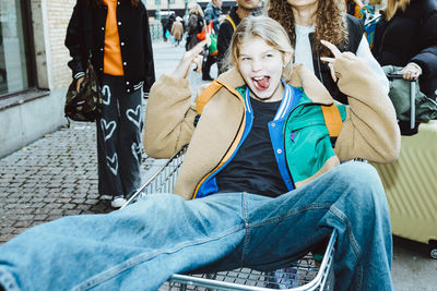 Girl sticking out tongue and gesturing horn sign sitting on shopping cart at footpath