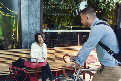 Businessman with bicycle talking to colleague sitting at sidewalk cafe