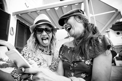 Low angle view of happy friends using smart phone while sitting against built structure