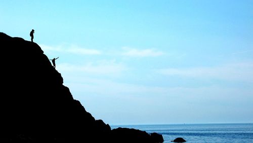Low angle view of silhouette men on cliff by sea against blue sky