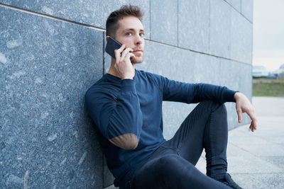 Side view of modern male in casual wear sitting near stone wall and br