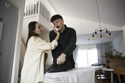 Woman holding smart phone on boyfriend's ear while ironing clothes at home