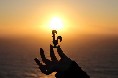 Cropped image of person holding silhouette rooster figurine by sea against sky during sunset