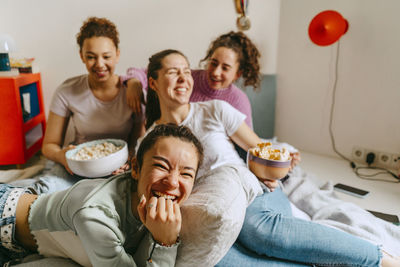 Cheerful female friends watching tv while enjoying snacks at home