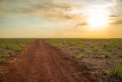 Dirt road amidst field against sky during sunset