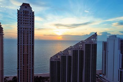 Modern buildings by sea against sky during sunset