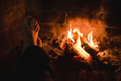 Crop anonymous person warming barefoot legs near yellow flames rising up from bonfire in nature at night