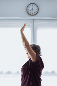 Side view of elderly female in activewear with short gray hair raising arms while doing stretching exercise against window in sunlit room in morning at home