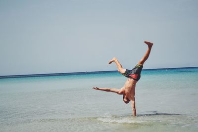 Full length of young shirtless man practicing on seashore against clear sky