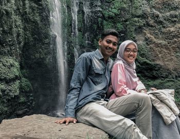 Portrait of couple smiling while sitting on rock against waterfall