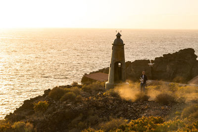 Man flying drone by beacon on hill against sea during sunset