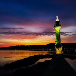 Lighthouse by silhouette building against sky during sunset