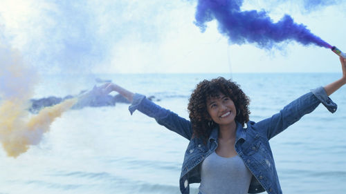 Smiling young woman with distress flare standing against sea