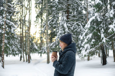 A nice middle-aged blonde woman walks through the winter forest with a cup of hot drink. 