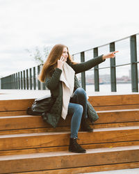 Side view of woman talking over smart phone while sitting on steps against sky