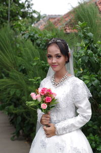 Portrait of beautiful bride holding flowers while standing against plants