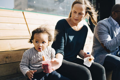 Woman sitting with man feeding drink to daughter while sitting at park