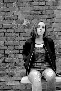 Portrait of young woman sitting on bench against brick wall