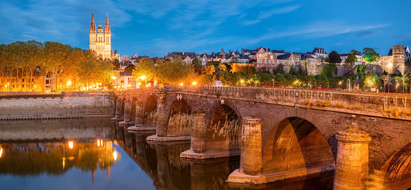 Illuminated arch bridge over river by buildings against sky in city