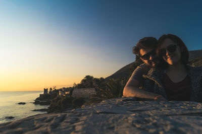 Young couple sitting on rock by sea against clear sky during sunset