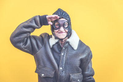 Portrait of senior woman wearing glasses while saluting against yellow background
