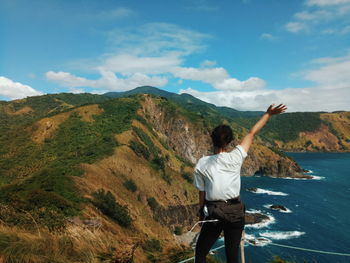 Rear view of woman waving hand while standing on cliff by sea against sky