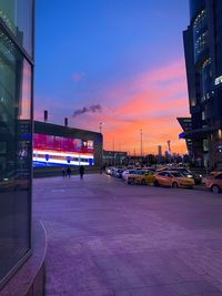 Cars on street by buildings against sky at sunset