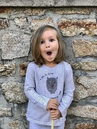 Three year old girl standing in front of the stone wall with a funny face 