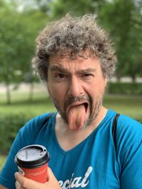 Portrait of a curly man in a blue t-shirt holding a paper cup with coffee and showing his tongue 