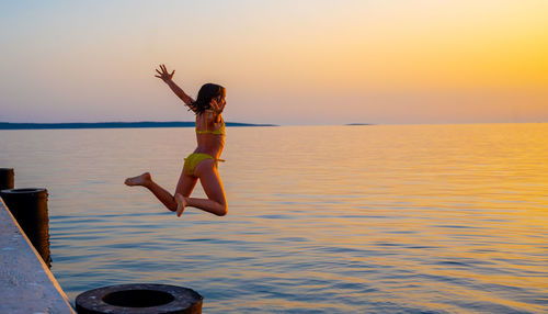 Girl with arms outstretched jumping in sea against sky during sunset