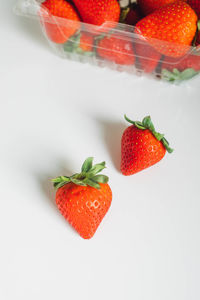 Two strawberries on white table with plastic carton of strawberries in the background