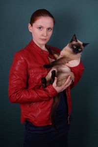 Portrait of young woman holding siamese cat against gray background