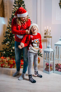 Happy mom and laughing daughter in christmas sweaters stand by the christmas tree and have fun
